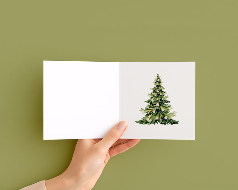 May include: A blank greeting card held by a hand, featuring a watercolor illustration of a green Christmas tree with white accents. The card is open, with the tree on the right side against a white background.