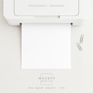 Puede incluir: Una impresora blanca con el texto "PHOTOSHOP • PHOTOPEA" se muestra, con un papel blanco en blanco que emerge. Dos clips plateados están a la derecha. La parte inferior de la imagen tiene el texto "MOCKUP HAUS CO" y "PSD SMART OBJECT • JPG".