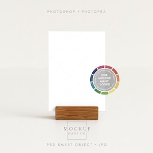 Puede incluir: Una tarjeta rectangular blanca se sostiene en un soporte de bloque de madera. Un gráfico de rueda de color con el texto "ONE MOCKUP, MANY LOOKS!" está a la derecha. También se ven las palabras "PHOTOSHOP PHOTOPEA" y "MOCKUP HAUS CO".