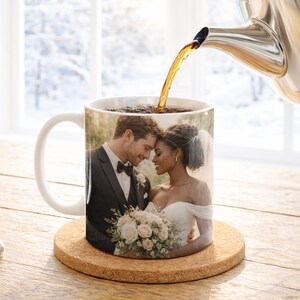 May include: A white ceramic mug featuring a wedding photo of a couple, with coffee being poured from a silver teapot. The mug sits on a round cork coaster. The background shows a snowy outdoor scene.
