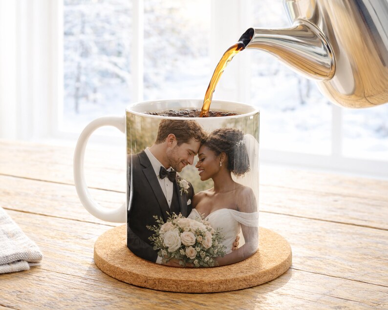 May include: A white ceramic mug featuring a wedding photo of a couple, with coffee being poured from a silver teapot. The mug sits on a round cork coaster. The background shows a snowy outdoor scene.
