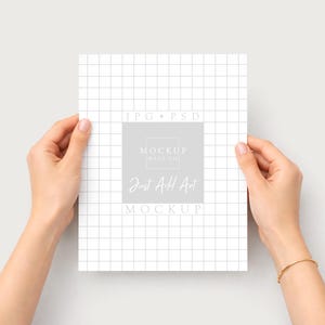 Puede incluir: Una maqueta de papel blanco con un patr&oacute;n de cuadr&iacute;cula es sostenida por dos manos. El papel tiene un cuadrado gris con el texto "MOCKUP HAUS CO" y "Just Add Art MOCKUP". Una pulsera dorada est&aacute; en la mu&ntilde;eca derecha.
