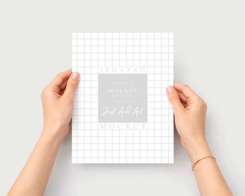Puede incluir: Una maqueta de papel blanco con un patr&oacute;n de cuadr&iacute;cula es sostenida por dos manos. El papel tiene un cuadrado gris con el texto "MOCKUP HAUS CO" y "Just Add Art MOCKUP". Una pulsera dorada est&aacute; en la mu&ntilde;eca derecha.