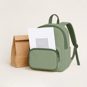 May include: A sage green backpack with a front pocket and a top handle. A brown paper lunch bag sits to the left of the backpack. A white notepad with grid lines is in the front pocket.
