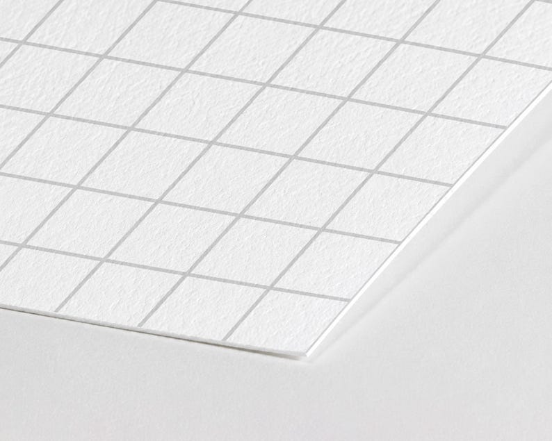May include: A sheet of white paper with a light gray grid pattern printed on the surface. The paper has a textured finish and is positioned at an angle, showing a sharp corner and straight edges against a plain white background.