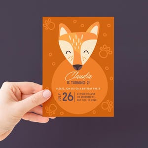 Peut inclure: Une invitation d'anniversaire orange avec une illustration de renard de dessin anim&eacute;. L'invitation indique "Claudia a 2 ans !" et comprend la date du 26 juillet, ainsi que les d&eacute;tails de la f&ecirc;te. Le design comprend des empreintes de pattes et des cercles.