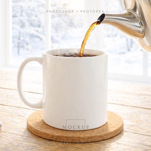 May include: A white ceramic mug is being filled with a dark liquid from a stainless steel teapot. The mug sits on a round cork coaster on a wooden table. The background shows a snowy winter scene through a window.