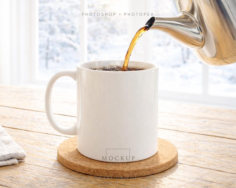 May include: A white ceramic mug is being filled with a dark liquid from a stainless steel teapot. The mug sits on a round cork coaster on a wooden table. The background shows a snowy winter scene through a window.