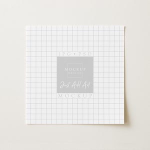 May include: Square white paper with a grid pattern, featuring the text "JPG PSD" at the top and "MOCKUP HAUS CO" and "Just Add Art MOCKUP" in a central gray square. The paper is on a light beige surface.