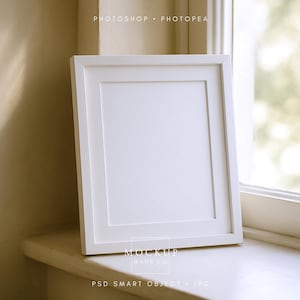 Puede incluir: Un marco de fotos cuadrado blanco con un paspartú blanco está apoyado en un alféizar blanco. El marco está inclinado ligeramente hacia el espectador, con el texto "PHOTOSHOP • PHOTOPEA" en la parte superior. El marco está frente a una ventana.
