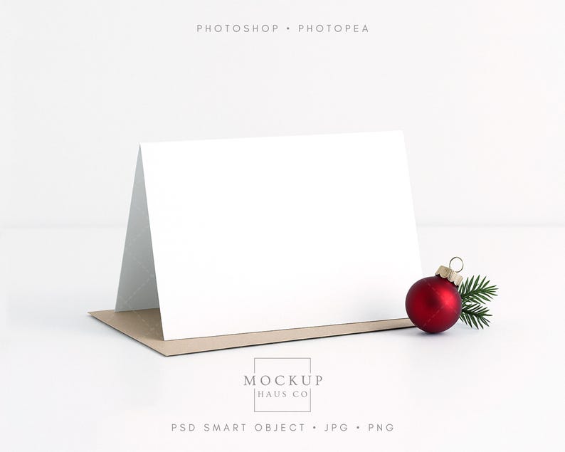 May include: A blank white greeting card with a folded design, resting on a brown card base. A red Christmas ornament with a gold cap and a sprig of green pine needles sits to the right. The text "PHOTOSHOP &bull; PHOTOPAE" is at the top.