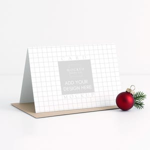 May include: A greeting card mockup with a grid pattern, featuring the text "Add Your Design Here." A red Christmas ornament and a sprig of evergreen are placed next to the card. The card is on a brown envelope.