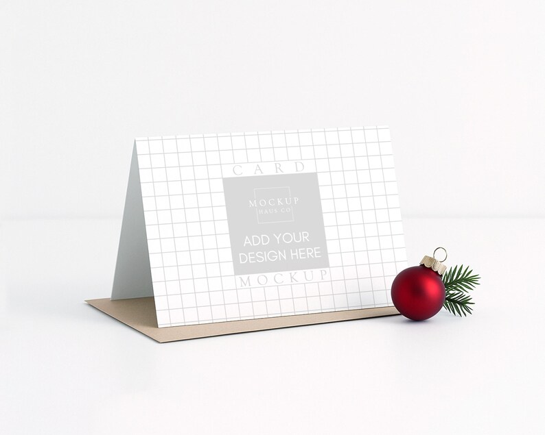 May include: A greeting card mockup with a grid pattern, featuring the text "Add Your Design Here." A red Christmas ornament and a sprig of evergreen are placed next to the card. The card is on a brown envelope.