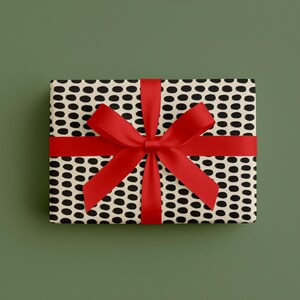 May include: A rectangular gift box wrapped in patterned paper with black oval shapes on a cream background. A bright red ribbon is tied around the box, forming a bow on top. The background is a solid, muted green.