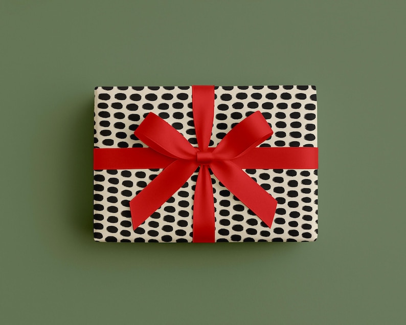 May include: A rectangular gift box wrapped in patterned paper with black oval shapes on a cream background. A bright red ribbon is tied around the box, forming a bow on top. The background is a solid, muted green.