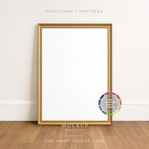 May include: A gold-framed rectangular picture frame leaning against a white wall on a wooden floor. The frame has a gold finish and a white interior. The image includes the text "PHOTOSHOP • PHOTOPEA" and "MOCKUP HAUS CO". A color wheel is in the lower right corner.
