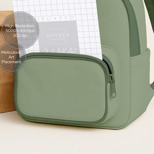 May include: A close-up of a sage green backpack with a front zippered pocket. A white paper with grid lines and text is partially inserted into the pocket. A brown paper bag is on the left. Text on the paper includes "High Resolution" and "Meticulous Art Placement".