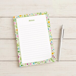May include: A floral-bordered notepad with the word "Notes" in green, and lined pages. A silver pen rests beside the notepad on a light wood surface. The notepad features a colorful floral design with pink, yellow, and blue flowers.