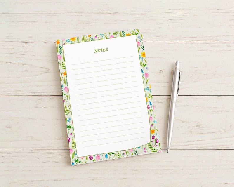 May include: A floral-bordered notepad with the word "Notes" in green, and lined pages. A silver pen rests beside the notepad on a light wood surface. The notepad features a colorful floral design with pink, yellow, and blue flowers.