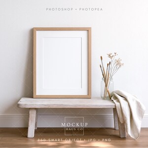May include: A wooden framed art print mockup with a white mat, resting on a distressed wooden bench. A glass jar with paintbrushes and dried flowers sits beside a folded cream-colored cloth. The background is a white wall and wooden floor.