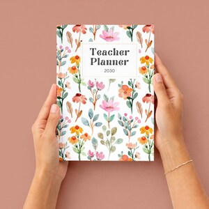 K&ouml;nnte beinhalten: Ein floraler Lehrerplaner f&uuml;r 2030, gehalten von H&auml;nden vor einem einfarbigen Hintergrund. Der Planer hat ein wei&szlig;es rechteckiges Textfeld mit den Worten "Teacher Planner 2030". Das Cover ist mit Aquarellblumen in Rosa, Orange und Gr&uuml;n verziert.