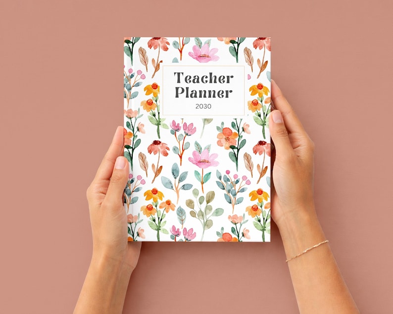 K&ouml;nnte beinhalten: Ein floraler Lehrerplaner f&uuml;r 2030, gehalten von H&auml;nden vor einem einfarbigen Hintergrund. Der Planer hat ein wei&szlig;es rechteckiges Textfeld mit den Worten "Teacher Planner 2030". Das Cover ist mit Aquarellblumen in Rosa, Orange und Gr&uuml;n verziert.