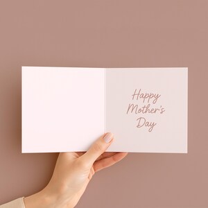 May include: A white greeting card with the text "Happy Mother's Day" in a cursive font. The card is held open, revealing the message, against a solid brown background.
