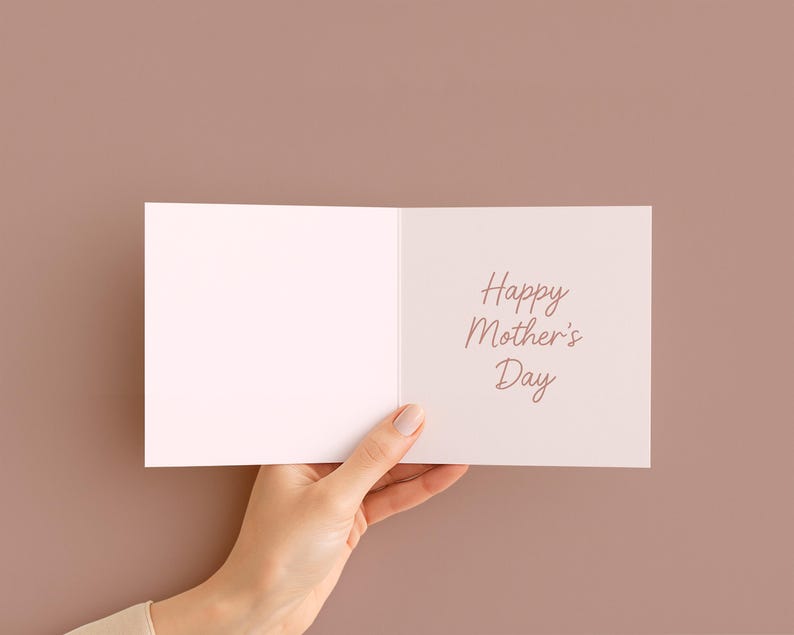 May include: A white greeting card with the text "Happy Mother's Day" in a cursive font. The card is held open, revealing the message, against a solid brown background.