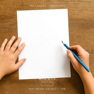 May include: A blank white paper with a subtle grid pattern is positioned on a wooden surface. A hand holds a blue pencil, poised to write or draw. The image includes the text "PHOTOSHOP • PHOTOPEA" and "MOCKUP HAUS CO".
