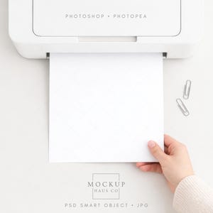 Puede incluir: Una impresora blanca con el texto "PHOTOSHOP • PHOTOPEA" en la parte superior. Se está imprimiendo un papel blanco en blanco, sostenido por una mano. Dos clips plateados están a la derecha. La parte inferior de la imagen tiene el texto "MOCKUP HAUS CO" y "PSD SMART OBJECT • JPG".