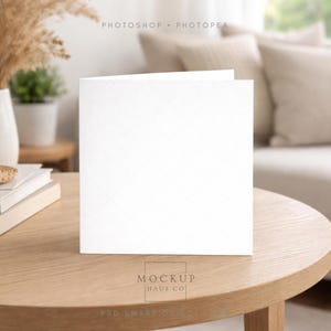 Pode incluir: Um cartão de felicitações branco em branco, com um padrão de diamante subtil, está aberto sobre uma mesa de madeira clara. A mesa está em frente a um sofá com almofadas de cor neutra. As palavras "MOCKUP HAUS CO" estão na parte inferior do cartão.