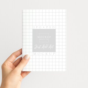 May include: A white notebook with a grid pattern is held by a hand. The notebook features a grey square with the text "MOCKUP HAUS CO" and "Just Add Art MOCKUP". The background is plain white.