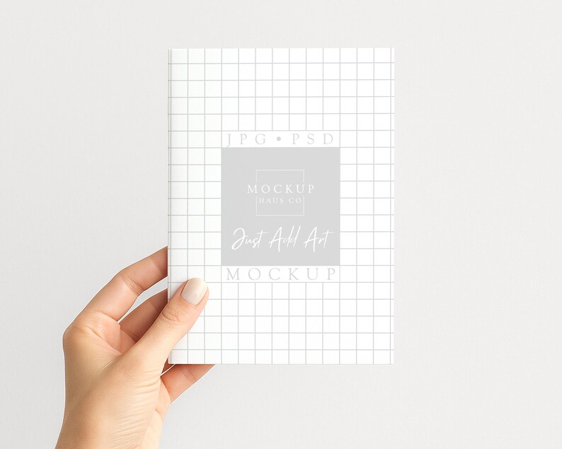 May include: A white notebook with a grid pattern is held by a hand. The notebook features a grey square with the text "MOCKUP HAUS CO" and "Just Add Art MOCKUP". The background is plain white.