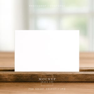 May include: A blank white card stands upright on a wooden surface, with a blurred background. The card is centered, offering space for text or design. The wooden base has a natural grain, and the scene is well-lit, suggesting a studio setting.