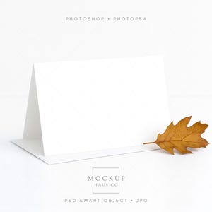 Pode incluir: Um cartão branco dobrado sobre uma superfície branca. À direita, uma folha de carvalho castanha e seca. O texto "PHOTOSHOP • PHOTOPAE" está no topo, e "MOCKUP HAUS CO" e "PSD SMART OBJECT • JPG" estão na parte inferior.