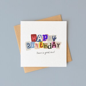 以下が含まれることがあります： カラフルな切り抜き文字で「HAPPY BIRTHDAY」と書かれた白いバースデーカード。メッセージ「Have a good one!」が下に印刷されています。カードは茶色の封筒の上にあり、水色の背景に配置されています。