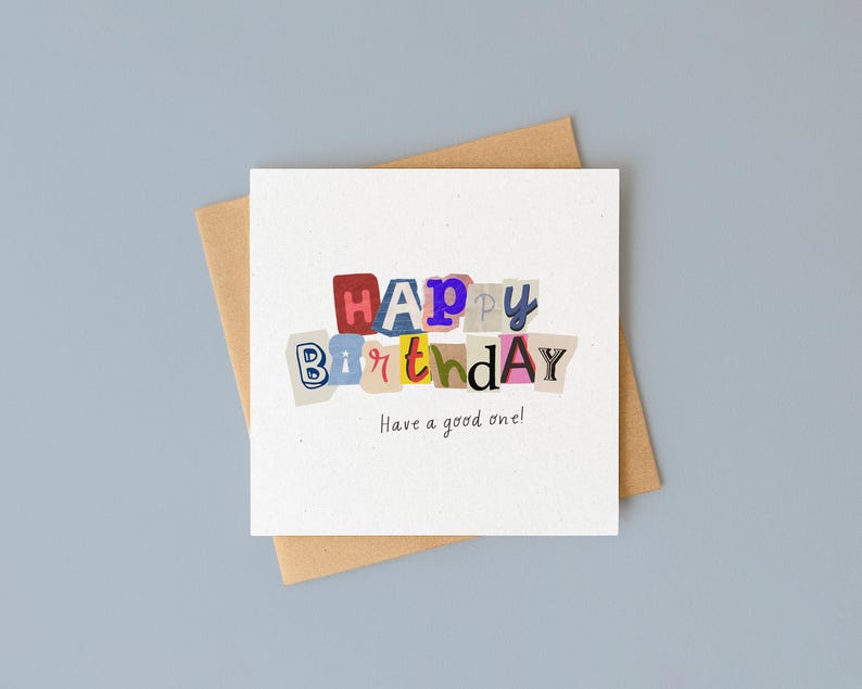 以下が含まれることがあります： カラフルな切り抜き文字で「HAPPY BIRTHDAY」と書かれた白いバースデーカード。メッセージ「Have a good one!」が下に印刷されています。カードは茶色の封筒の上にあり、水色の背景に配置されています。