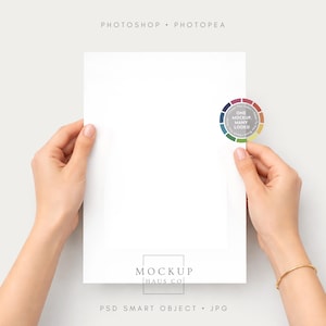 Op de afbeelding: Een wit papier wordt door twee handen vastgehouden, met een gouden armband om een pols. Een kleurrijk cirkelvormig logo is aan het papier bevestigd. De woorden "Photoshop Photpea" staan bovenaan, en "Mockup Haus Co" en "PSD Smart Object JPG" onderaan.