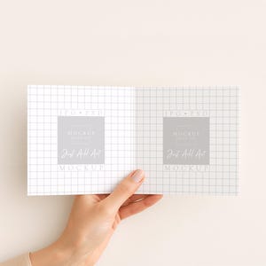 May include: A hand holding an open notebook with a grid pattern. The notebook has a gray square with the text "MOCKUP" and "Just Add Art" printed on it. The background is a solid, light beige color.