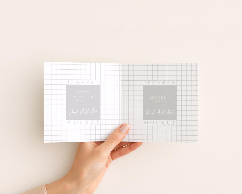 May include: A hand holding an open notebook with a grid pattern. The notebook has a gray square with the text "MOCKUP" and "Just Add Art" printed on it. The background is a solid, light beige color.