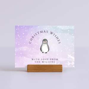 May include: A Christmas card with a watercolor background in shades of purple and blue, featuring a penguin illustration. The card reads "Christmas Wishes" and "With Love From The Wilsons". The card is held by a wooden block.
