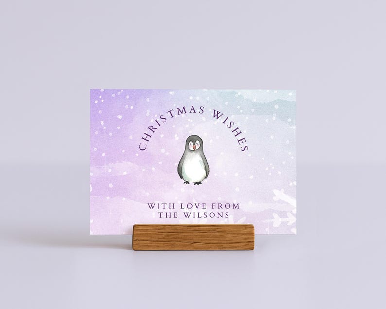 May include: A Christmas card with a watercolor background in shades of purple and blue, featuring a penguin illustration. The card reads "Christmas Wishes" and "With Love From The Wilsons". The card is held by a wooden block.