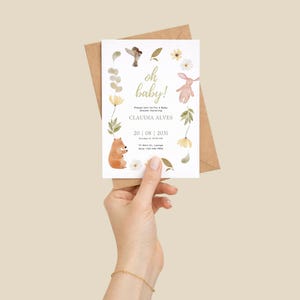 May include: A baby shower invitation with the words "oh baby!" in gold script. The invitation features watercolor illustrations of a bear, bunny, bird, and flowers. The invitation is held in front of a brown envelope.