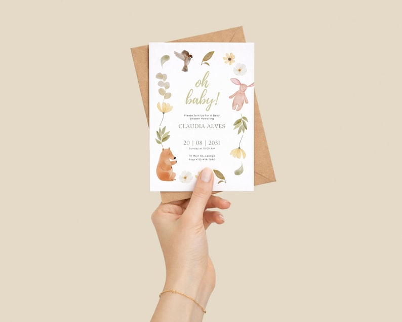 May include: A baby shower invitation with the words "oh baby!" in gold script. The invitation features watercolor illustrations of a bear, bunny, bird, and flowers. The invitation is held in front of a brown envelope.