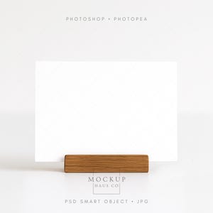 Könnte beinhalten: Eine weiße rechteckige Karte wird von einem rechteckigen Holzhalter gehalten. Der Holzhalter ist hellbraun. Das Bild enthält den Text "MOCKUP HAUS CO" und "PSD SMART OBJECT . JPG".