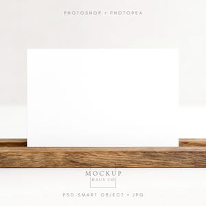 Pode incluir: Um cartão retangular branco em branco está em uma base de madeira rústica. As palavras "PHOTOSHOP • PHOTOPAE" estão no topo, e "MOCKUP HAUS CO" e "PSD SMART OBJECT • JPG" na parte inferior. Fundo branco.