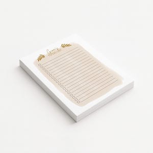 Peut inclure: Une pile de blocs-notes blancs avec un lavis d'aquarelle beige et du papier lign&eacute;. Le bloc-notes porte l'inscription "Grind's To Do List" en lettres dor&eacute;es avec des accents de feuilles d'or.
