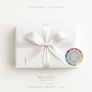 以下が含まれることがあります： 白い長方形のギフトボックスで、サテンのリボンと蝶結びで結ばれています。ボックスは白い紙で包まれています。「ONE MOCKUP, MANY LOOKS!」というテキストが入った円形のグラフィックが右側にあります。「MOCKUP HAUS CO」というテキストが下にあります。