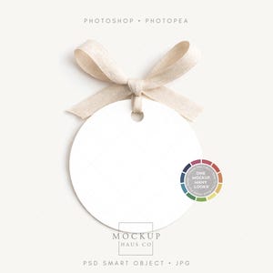 May include: A blank, white, circular tag with a beige ribbon bow. The tag has a hole for hanging. The image includes the text "Photoshop + Photopea" and "Mockup Haus Co."