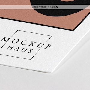 May include: A white paper mockup featuring a textured surface with a black border and a dusty rose colored section. The text "ADD YOUR DESIGN" is printed in black at the top, and the logo "MOCKUP HAUS" is enclosed in a thin black square.